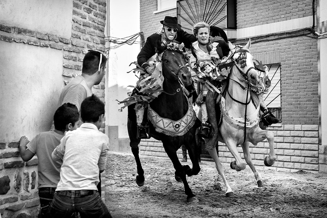 Two riders between buildings, watched by a group of people.